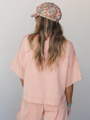 Shot from behind, a model wears a soft blush-coral matching set — boxy tee and wide-leg pants — with visible contrast stitching throughout, topped with a floral embroidered baseball cap and a turquoise beaded bracelet peeking out at the wrist.