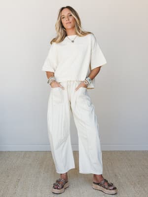 A cream matching set — oversized boxy tee and wide-leg pants — styled with stacked beaded bracelets, a turquoise pendant necklace, and platform espadrille sandals against a clean studio wall, giving off that effortlessly relaxed, nowhere-to-be energy