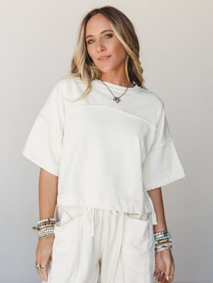 A cream matching set — oversized boxy tee and relaxed drawstring pants — styled with stacked beaded bracelets and a turquoise pendant necklace, shot against a soft neutral backdrop with an easy, effortless feel.