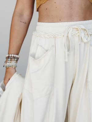 A close-up waist shot of cream wide-leg pants with a smocked elastic waistband and drawstring tie, styled with a glimpse of a golden yellow crop top and a stack of beaded bracelets — relaxed and sun-warmed, like a slow morning somewhere coastal.