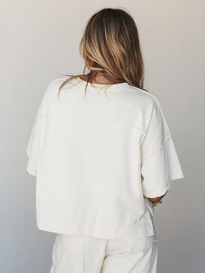 Shot from behind, a model wears a cream oversized cropped sweatshirt with visible seam detailing paired with matching cream wide-leg pants — the whole look reads like a soft, intentional set you'd reach for on a slow Saturday.