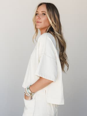 A model shot in soft studio light wearing a cream oversized short-sleeve top with visible seam detailing, paired with matching cream wide-leg pants — the whole look has that effortless, lived-in ease, accessorized with a stack of beaded bracelets tha