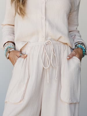 A close-up shot of a cream gauze co-ord set — button-front top meets drawstring wide-leg pants with patch pockets, both hands tucked in, stacked turquoise and beaded bracelets adding just the right amount of personality.