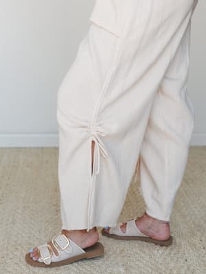A close-up of wide-leg cream textured pants with a ruched drawstring hem detail, paired with double-buckle slide sandals on a woven rug — easy, intentional, and the kind of outfit that looks effortless but isn't accidental.