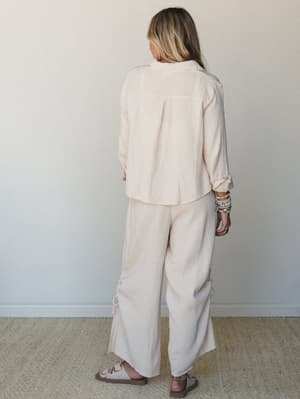A breezy matching set in soft blush-cream gauze — wide-leg pants with a side tie detail paired with a relaxed button-down top, shot from behind with layered bead bracelets adding just enough personality.