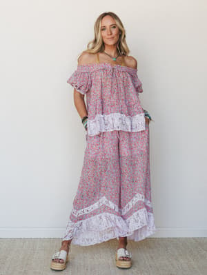 A matching floral two-piece set in soft lilac and coral — off-shoulder top with smocked trim and lace ruffle hem paired with wide-leg pants finished in lace border detail, styled with stacked bracelets and platform sandals against a clean white wall.