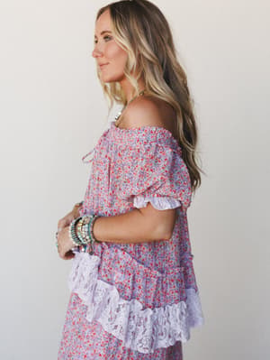 A side-profile shot of a model in a flowy lilac ditsy floral off-shoulder dress with lace ruffle trim, styled with a stack of beaded bracelets — romantic and free-spirited with a soft, lived-in ease.