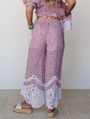 A back-facing model wears a matching floral two-piece set — cropped top and wide-leg pants with dramatic lace hem panels — paired with platform espadrille sandals and layered bracelets, shot against a soft neutral studio wall.
