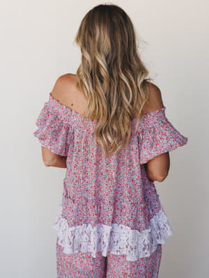 A back-facing shot of a flowy off-shoulder top in a ditsy floral print — lavender and red — finished with a lace ruffle hem that makes it feel like something you'd find at a vintage market and never stop wearing.
