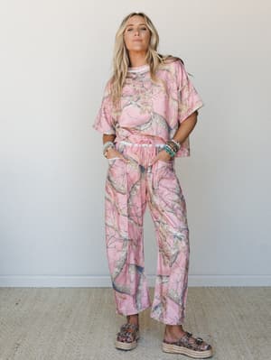 A matching pink camo two-piece set — oversized tee and wide-leg drawstring pants — styled with platform espadrille sandals and a stack of beaded bracelets, shot against a clean neutral wall with a relaxed, confident ease.
