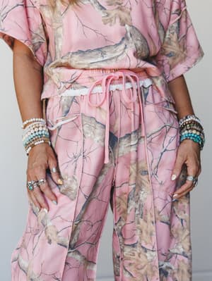 A close-up torso shot of a pink camo matching set — cropped top and wide-leg drawstring pants — styled with layered beaded bracelets, turquoise jewelry, and silver rings for a playful, accessory-heavy look.