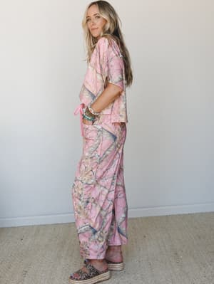 A matching pink camo two-piece set — cropped top and wide-leg maxi pants — styled with platform espadrille sandals and a stacked bracelet arm party, shot in profile against a clean white wall with an easy, relaxed confidence.