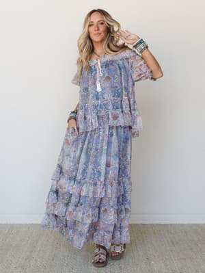 A flowy, tiered floral maxi dress in soft periwinkle and blush tones styled with stacked beaded bracelets and flat sandals — the kind of outfit that feels equally at home at a summer wedding or wandering a farmers market.