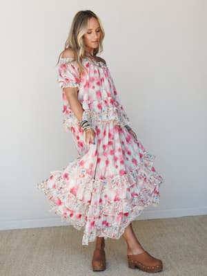 A flowy floral maxi dress in soft cream and pink moves mid-twirl, styled with stacked bracelets and wood-platform clogs for a free-spirited, romantic feel.