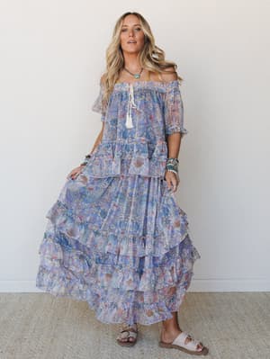 A flowy, tiered maxi dress in a dreamy blue and pink floral paisley print, worn off-the-shoulder with layered boho jewelry and flat sandals — the kind of outfit that looks equally at home at a beach wedding or wandering a farmers market.