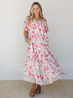 A flowy, tiered floral maxi dress in blush and hot pink worn off-the-shoulder with brown clogs and stacked beaded bracelets — effortlessly romantic and made for spinning in.