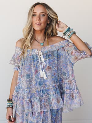 A flowy off-shoulder top in a dreamy blue and pink floral paisley print, styled with stacked boho bracelets and a turquoise pendant — the kind of outfit that looks like it was made for wandering somewhere beautiful.