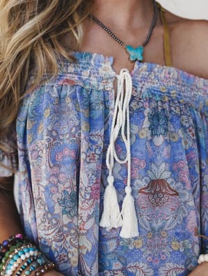 A close-up lifestyle shot of a flowy boho top in a dreamy periwinkle and pink floral print, styled with tassel tie detail at the neckline, layered bead bracelets, and a turquoise butterfly pendant — the kind of outfit that looks like it came from a r