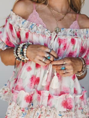 A close-up torso shot showcasing a flowy floral dress with cold-shoulder smocking and tassel ties, styled with layered beaded bracelets, stacked silver rings with turquoise stones, and a delicate layered gold necklace — the whole thing feels like a S