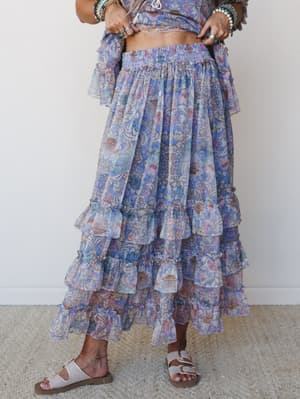 A flowy tiered maxi skirt in a dreamy blue and blush floral print, styled as a matching set with a cropped top, stacked boho jewelry, and flat buckle sandals — the kind of outfit that makes a farmers market feel like an event.
