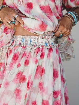 A close-up waist shot of a flowy floral skirt set in watercolor pink roses, styled with layered silver and turquoise rings and stacked bracelets — the kind of outfit that makes you want to spin.
