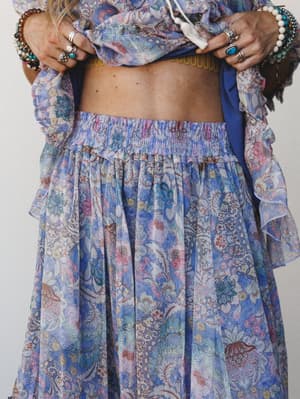 A close-cropped torso shot of a flowy floral maxi skirt with a smocked waist, styled as a set with a matching ruffle-sleeved top — her hands rest at her waist, stacked with silver rings and beaded bracelets, the whole look landing somewhere between a