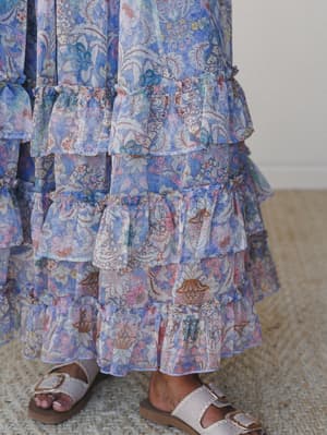 A close-up of a dreamy tiered maxi skirt in a layered botanical print — all soft periwinkle, blush, and warm rust — paired with casual buckle sandals on a jute rug, giving off easy summer energy.