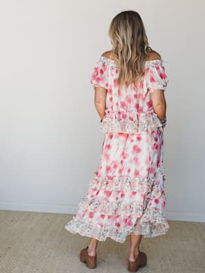 A flowy floral skirt set in pink and cream photographed from behind, with off-shoulder ruffle top and tiered maxi skirt that feels like a garden party waiting to happen — paired with wooden clogs for that perfect lived-in boho finish.