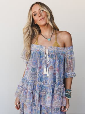 A flowy off-shoulder floral top — part of a matching set — in soft periwinkle and blush with tassel tie detail, styled with a visible golden lace bralette, turquoise pendant necklace, and stacked beaded bracelets for a free-spirited, lived-in boho fe