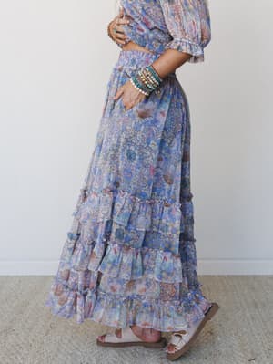 A side-profile shot of a flowy floral maxi skirt set in soft periwinkle and blush, styled with stacked beaded bracelets and flat sandals — the kind of outfit that feels like a deep breath.
