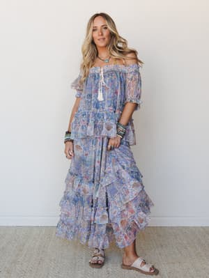 A flowy, floral-printed off-shoulder maxi set in dreamy periwinkle blue and blush tones, styled with beaded bracelets, a turquoise necklace, and flat sandals — the kind of outfit that looks equally at home at a garden party or a sunset stroll.