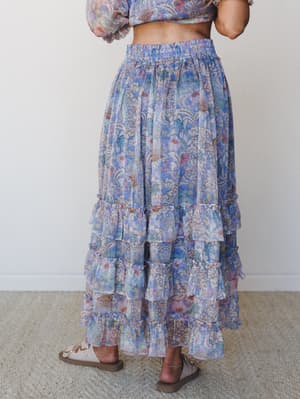 A back-view shot of a flowy tiered maxi skirt in a watercolor floral print — periwinkle blues, soft pinks, and dusty rose swirl together in a dreamy botanical pattern, styled as a matching set with a cropped long-sleeve top and neutral slide sandals.
