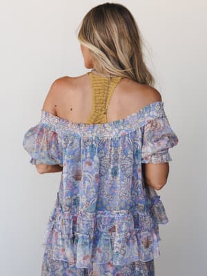 Shot from behind, a model wears a dreamy off-shoulder floral top with tiered ruffle skirt in soft periwinkle and blush — layered over a mustard crochet bralette that peeks out like a happy accident.