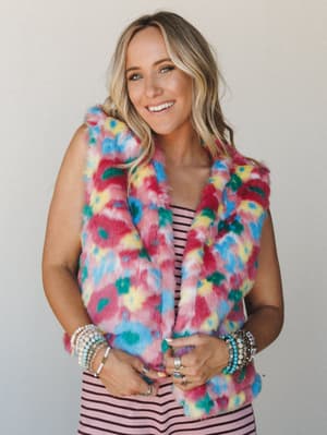 A multicolor faux fur vest in a bold floral patchwork print layered over a pink stripe top, styled with stacked beaded bracelets and silver rings — playful and full of personality against a simple light gray backdrop.