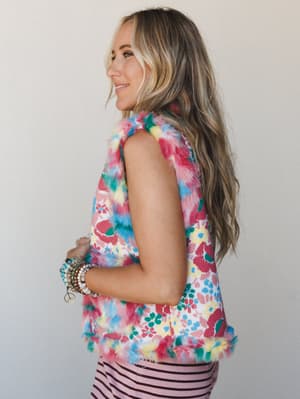 A model shot in profile wearing a sleeveless floral vest with multicolor faux fur trim at the collar, armholes, and hem — playful and a little maximalist, styled with a pink striped skirt and a stack of beaded bracelets.
