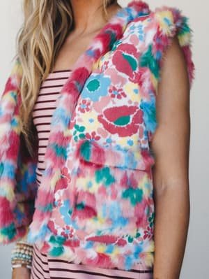A close-up torso shot of a multicolor faux fur floral vest layered over a pink stripe knit top — the kind of outfit that commits fully to joy and somehow makes it work.