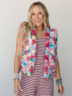 A bright, joyful shot of a model layering a multicolor floral faux fur vest over a blush pink striped set — the kind of outfit that makes a statement without trying too hard, stacked bracelets and all.