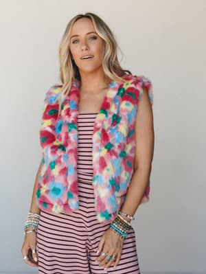 A multicolor faux fur vest layered over a pink striped set, styled with stacked bracelets and rings — the kind of outfit that commits fully to the fun and somehow pulls it off.