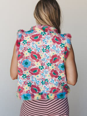 A back-view shot of a cropped floral knit vest with multicolor faux fur trim along the edges, layered over a pink striped bottom — playful and a little unexpected in the best way.