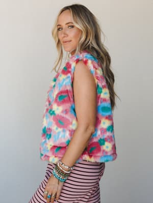 A multicolor faux fur floral vest worn over a pink striped bottom, styled with stacked beaded bracelets and rings — the kind of outfit that has a personality before you even walk in the room.