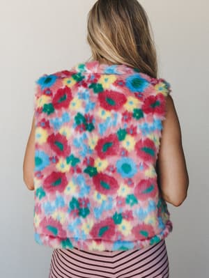 A back-view shot of a cropped faux fur vest in a bold multicolor floral print — the kind of piece that makes a statement without saying a word — styled over a pink striped bottom against a clean neutral wall.