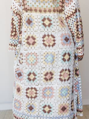 A close-up torso shot of a long crochet duster covered in granny square motifs in a soft pastel patchwork — the kind of piece that looks like someone's most loved summer memory made wearable.