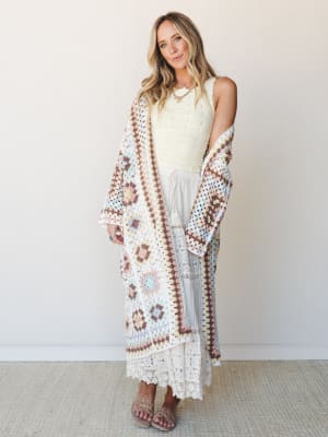 A relaxed full-body shot of a model layering a granny-square crochet duster in soft pastels over a cream pointelle tank and a white lace maxi skirt — the whole look has that effortless, serotonin-boosting boho energy.