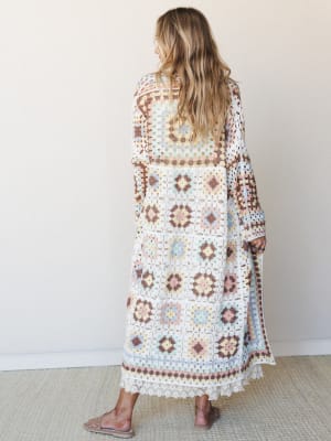 Shot from behind, a model in a floor-length granny square crochet duster in soft pastel tones stands against a warm white wall — the kind of piece that looks like it was made from a really good vintage find.