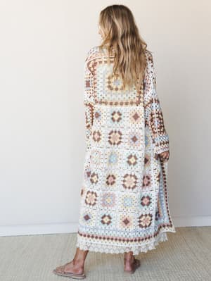 Shot from behind, a model in a floor-length granny square crochet duster in soft pastel tones stands against a warm white wall — the kind of piece that looks like it was made from a really good vintage find.