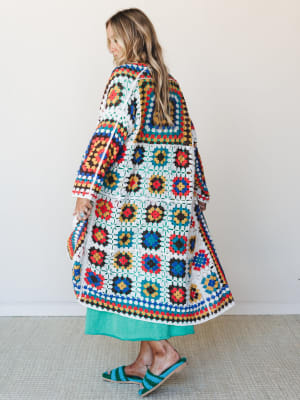 A model shot from behind showing a sweeping granny square crochet duster in bold multicolor layered over a kelly green midi skirt, styled with matching crochet slide sandals — the whole look has that 'found it at a vintage market and never looked bac