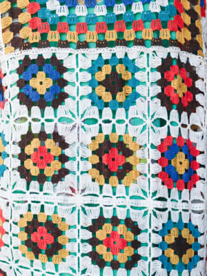 A close-up detail shot of a multicolor crochet duster covered in granny square motifs — layered flowers in red, gold, blue, and brown connected by an open white crochet lattice, with a green lining visible beneath.