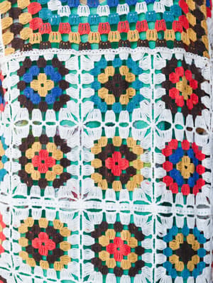 A close-up detail shot of a multicolor crochet duster covered in granny square motifs — layered flowers in red, gold, blue, and brown connected by an open white crochet lattice, with a green lining visible beneath.