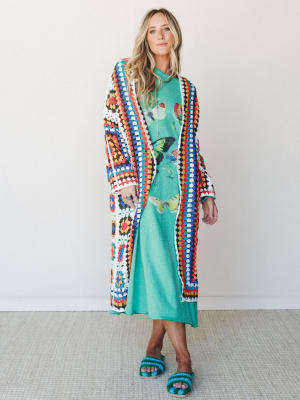 A floor-grazing granny square crochet duster in vivid multicolor layered over a mint green butterfly graphic midi dress, styled with matching crochet sandals — the kind of outfit that looks like it was assembled with pure joy.