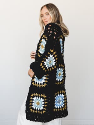 A granny square crochet cardigan in black with pops of dusty blue, golden mustard, and cream — worn over white pants against a clean studio wall, with that easy 'I've had this forever and it goes with everything' energy.